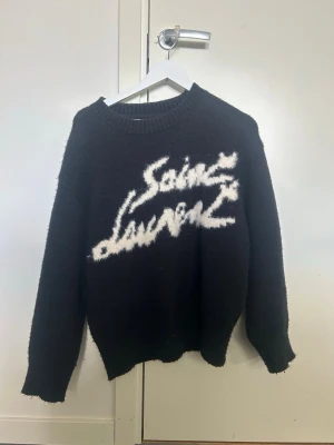 Svart Saint Laurent stickad tröja - Tvärfet svart stickad tröja från Saint Laurent med vit, fluffig logotyp framtill. Tröjan har rund halsringning, långa ärmar och ribbstickade muddar. Perfekt för dig som gillar streetstyle och vill sticka ut med en exklusiv look.
