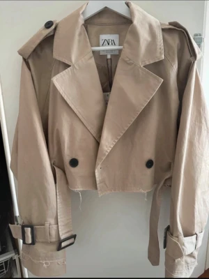 Beige cropped trenchcoat från Zara - Trendig beige cropped trenchcoat från Zara med stora slag, svarta knappar och bältesdetaljer vid ärmsluten. Jackan har axelklaffar och en avslappnad passform som ger en cool look. Perfekt för dig som vill ha en modern twist på klassikern. I storlek M men passar mig som har S perfekt 