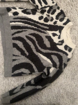 Stickad tröja med mönster - Cool stickad tröja med svart, grått och vitt djurmönster. Tröjan har rund halsringning, ribbade muddar och är långärmad. Perfekt för dig som gillar att sticka ut med ett unikt mönster och vill ha något både bekvämt och trendigt.