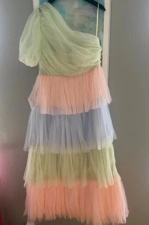 By malina Pastellfärgad tyllklänning - Superfin långklänning i pastellfärgad tyll med volanger och oneshoulder-design. Klänningen har lager i ljusgrön, ljusrosa och ljusblå nyans och ger en riktigt drömmig känsla. Perfekt för dig som vill sticka ut med en unik och färgglad look. Storlek 42- använd för fotografering, nyskick 