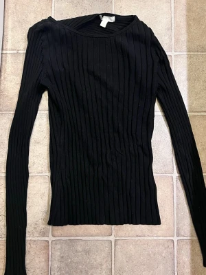 Svart ribbad långärmad topp - En svart långärmad topp med ribbad struktur och rund hals. Toppens material är stretchigt och figurnära, perfekt för att skapa en clean och stilren look. Passar dig som gillar minimalistiska plagg med enkel design.
