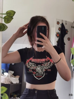 Svart croppad t-shirt med tryck - Cool svart croppad t-shirt med rött och vitt tryck framtill, motiv med örn och text. T-shirten har rund halsringning och korta ärmar. Perfekt för en edgy och avslappnad stil.