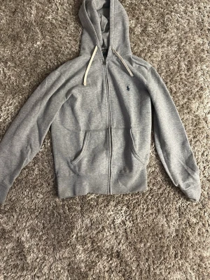 Grå hoodie från Polo Ralph Lauren - Snygg grå hoodie från Polo Ralph Lauren med dragkedja, huva med vita snören och klassisk blå logga broderad på bröstet. Tillverkad i mjukt bomullsmaterial och har två fickor framtill. Perfekt för en avslappnad och stilren look.
