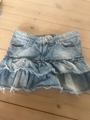 Jeanskjol med volanger från Gina Tricot - Snygg kort jeanskjol från Gina Tricot i blå denim med volangdetaljer och råa kanter. Kjolen har klassisk femficksdesign, bälteshällor och knappstängning framtill. Perfekt för dig som gillar en cool och avslappnad stil.