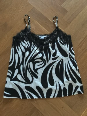 Svartvit linnetopp med spetsdetaljer - Snygg linnetopp med svartvitt abstrakt mönster och tunna axelband. Toppen har spetsdetaljer längs urringningen som ger en extra touch. Perfekt att styla till jeans eller kjol för en trendig look.