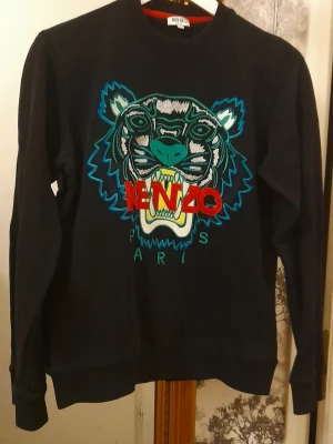 Svart Kenzo sweatshirt med tigertryck - Cool svart sweatshirt från Kenzo med ikoniskt broderat tigerhuvud i blått, grönt och vitt samt röd Kenzo-logga framtill. Tröjan har rund hals, långa ärmar och ribbade muddar.  Bra begagnat. Ser ny ut. Aningen stel pga tvätt. 