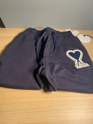 Mörkblå mjukisshorts Ami Paris - Mörkblå mjukishorts  från Ami Paris med elastisk midja och dragsko. Byxorna har en stor vit broderad Ami-hjärtlogga på ena benet. Perfekta för en chill och avslappnad stil. Tillverkade i mjukt material för extra komfort.