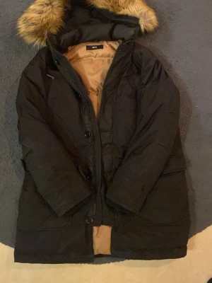 Svart dunjacka med fuskpäls från BOSS - Stilren svart dunjacka från BOSS med beige innerfoder och huva med fuskpäls. Jackan är fylld med 80% ankdun och 20% ankfjäder, perfekt för kalla dagar. Klassisk lång modell med dragkedja och knappar framtill.