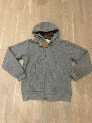 Grå zip-hoodie från Burberry - Snygg grå hoodie från Burberry med dragkedja, huva med klassiskt rutigt foder och dragsnören. Broderad logga på bröstet och två fickor framtill. Perfekt för en stilren och avslappnad look.