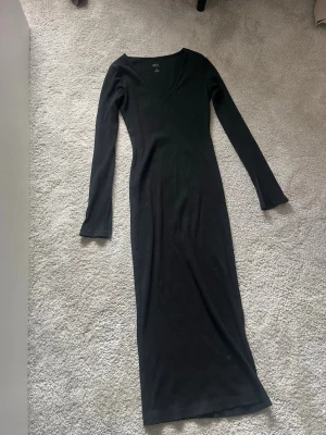 Svart ribbad långklänning - Svart långklänning (går strax under knäna) i ribbad bomullsmix. Klänningen har lång ärm med liten slit och v-ringning, sitter figurnära och är perfekt för en stilren look. Enkel men snygg design som passar till många olika tillfällen.