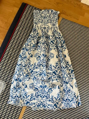Blåvit blommig sommarklänning - Supersöt blåvit klänning med blommigt mönster och smock upptill. Klänningen är ärmlös och har en luftig, lång modell som passar perfekt för varma dagar. Materialet känns lätt och bekvämt, och volangdetaljen upptill ger en extra charmig touch.