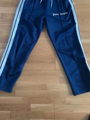 Blåa mjukisbyxor Palm Angels - Snygga blåa mjukisbyxor från Palm Angels med vita sidoränder och dragsko i midjan. Byxorna har en avslappnad passform och loggan broderad framtill. Perfekta för en trendig och sportig look.
