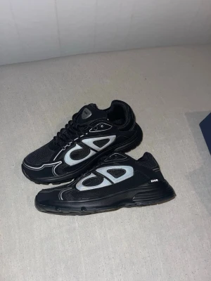 Christian Dior B30 Trainers  - Säljer ett par svarta Dior sneakers med tydliga silverfärgade detaljer och logga på sidan. Skorna har en chunky sula, mesh- och läderpaneler samt snörning framtill. Perfekta för dig som vill sticka ut med en lyxig och modern look. Finns kvitto.