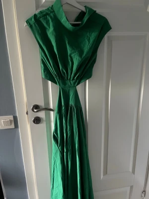 Grön långklänning med cut out - Snygg grön långklänning med markerad midja och cut out-detalj i sidan. Klänningen har ärmlös design och ett luftigt fall, perfekt för varma dagar. Tillverkad i ett lätt bomullstyg som känns bekvämt mot huden.