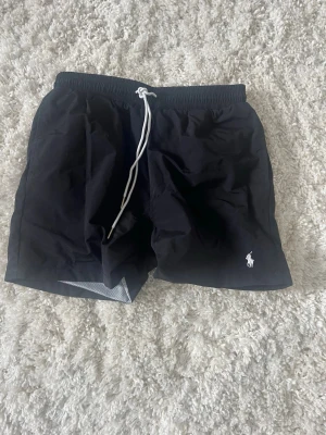 Svarta badbyxor från Polo Ralph Lauren - Svarta badbyxor från Polo Ralph Lauren med vit dragsko i midjan och klassisk logga broderad framtill. Innerbyxa i mesh för extra komfort. Perfekta för stranden eller poolen. Skriv vid frågor eller funderingar!