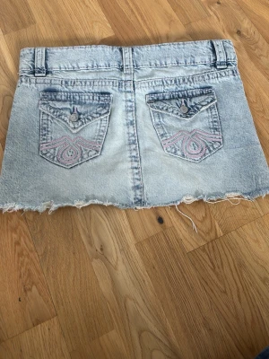 Ljusblå jeanskjol  - Snygg ljusblå jeanskjol med rå, fransig kant och slitna detaljer. Kjolen har två bakfickor med broderi och knapp, samt bälteshällor och klassisk jeansknäppning fram. Perfekt för en avslappnad och trendig look.