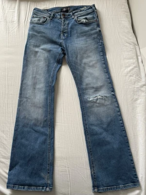 Blå bootcut jeans med slitningar - Bootcut jeans i mellanblå denim med slitningar på knät och fransade benslut. Klassisk femficksmodell med dragkedjegylf och lätt tvättad look. Perfekta för en avslappnad och trendig stil.