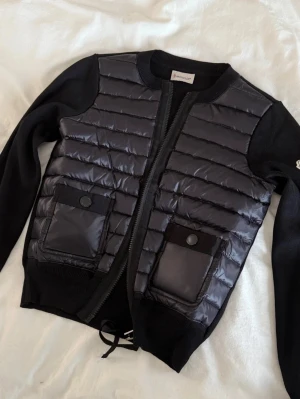 Svart pufferjacka från Moncler - Snygg svart pufferjacka från Moncler med quiltad front och två stora fickor med lock. Jackan har stickade ärmar och sidor, rund halsringning och dragkedja framtill. Perfekt för kalla dagar och riktigt trendig stil.