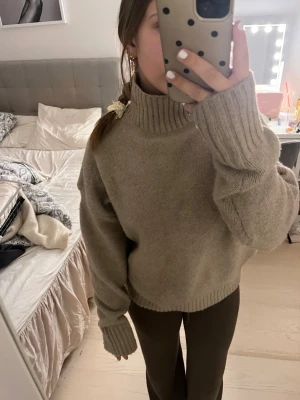 Beige stickad polotröja - Mysig beige stickad polotröja med oversized passform och långa ärmar. Tröjan har en hög ribbad krage och ribbade muddar vid ärmslut och nederkant. Perfekt för kalla dagar och enkel att matcha med olika outfits.