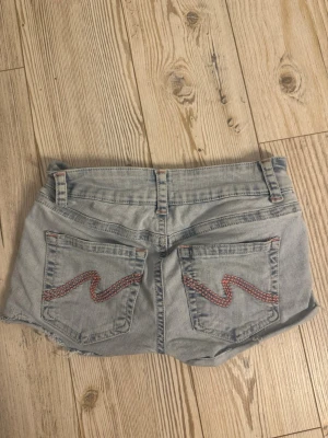 Ljusblå jeansshorts  - Snygga ljusblå jeansshorts med råa kanter och rosa broderade detaljer på bakfickorna. Inga tecken på användning 
