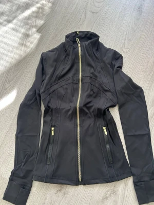 Svart Lulumelon sportjacka med guldiga dragkedjor - Snygg svart Lulumelon sportjacka med figurnära passform och guldiga dragkedjor framtill och vid fickorna. Jackan har hög krage och långa ärmar, perfekt för träning eller promenader. Tillverkad i stretchigt funktionsmaterial för extra komfort.