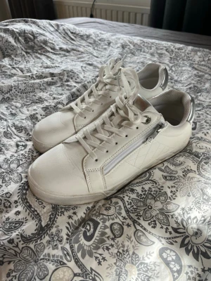 Vita sneakers med dragkedja - Snygga vita sneakers med silverdetaljer och dragkedja på sidan. Skorna har klassisk låg modell med vita skosnören och diskret mönster på sidan. Perfekta för en clean och stilren look. Materialet är troligtvis syntet eller läderimitation.Är använda en gång