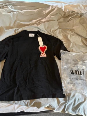 Svart t-shirt från Ami Paris - Svart t-shirt från Ami Paris med stort rött hjärta och A-logga på bröstet. Klassisk passform med rund hals och korta ärmar. Tillverkad i mjuk bomull, perfekt för en stilren och trendig look.
