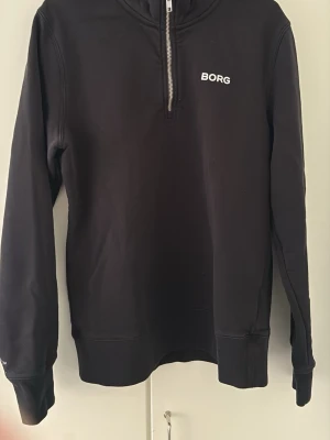 Svart halfzip hoodie från BORG - Svart hoodie från BORG med halv dragkedja framtill och vit logga på bröstet. Tröjan har långa ärmar, ribbade muddar och en stilren design. Perfekt för en avslappnad och sportig look.