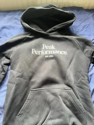 Svart Peak Performance hoodie - Svart hoodie från Peak Performance med vit logga och text 'Est. 1986' på bröstet. Klassisk känguruficka framtill och stor huva. Perfekt för en avslappnad och sportig stil. Mjuk insida och ribbade muddar vid ärmslut och nederkant.