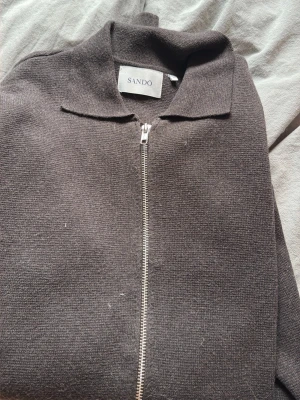 Brun ziptröja från Sandö - Mörkbrun stickad ziptröja från Sandö med klassisk krage och hel dragkedja framtill. Tröjan har en enkel och stilren design, perfekt för lager-på-lager och chill vardagsstil.