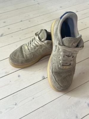 Nike Air Force 1 grå mocka sneakers - Nike Air Force 1 sneakers i grå mocka med klassisk siluett och beige sula. Skorna har perforerade detaljer på tån, vita skosnören och metallbricka med 'AF1' på snörningen. Perfekt för dig som gillar streetwear och tidlös design.