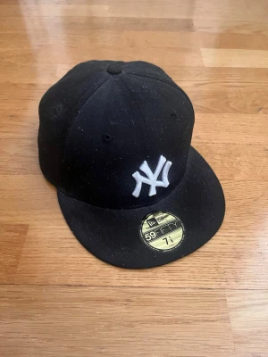 Svart New Era Yankees keps 59FIFTY - Säljer en svart New Era 59FIFTY keps med klassiskt New York Yankees-broderi i vitt framtill. Flat skärm och guldfärgad storleksdekal kvar på skärmen. Snygg streetwear-look och perfekt för dig som gillar sportiga accessoarer.