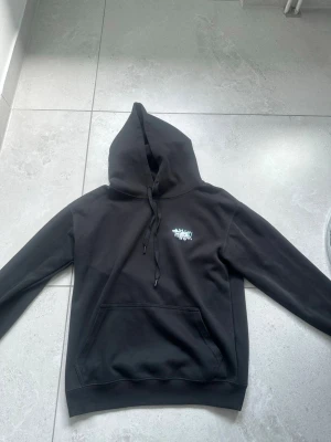 Svart hoodie från Amiri med tryck - Säljer en svart hoodie från Amiri med stor blommig print och AMIRI-text i turkost på ryggen samt mindre logga på bröstet. Hoodien har huva med dragsko och en stor magficka. Perfekt för dig som gillar streetwear och vill sticka ut lite extra.