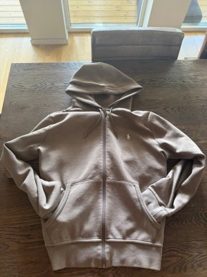 Beige hoodie med dragkedja från Polo Ralph Lauren - Snygg beige hoodie från Polo Ralph Lauren med dragkedja framtill och två stora fickor. Klassisk huva med dragsko och den ikoniska lilla loggan broderad på bröstet. Perfekt för en chill och avslappnad stil. Tillverkad i mjukt sweatshirtmaterial.