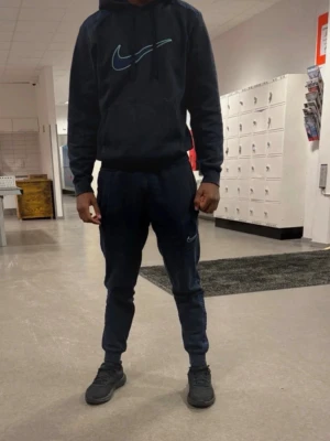 Svart Nike träningsset med orange detaljer - Snyggt svart träningsset från Nike med quiltad bröstpanel och halv dragkedja på tröjan. Både tröja och byxa har orange Nike-logga. Perfekt för träning eller chill. Materialet är mjukt och stretchigt för bästa komfort. 