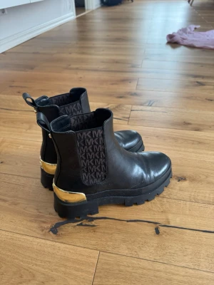 Svarta Chelsea boots från Michael Kors - Snygga svarta Chelsea boots i läder från Michael Kors med grov sula och gulddetaljer vid hälen. Resårpartierna har MK-logga i svart och mörkbrunt. Perfekta boots för dig som vill ha en trendig och lyxig look.