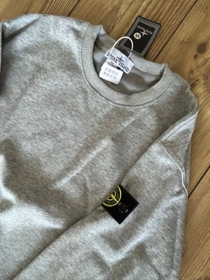 Grå sweatshirt från Stone Island - Snygg grå sweatshirt från Stone Island med klassisk rund halsringning och ikonisk logopatch på vänster ärm. Tillverkad i mjuk bomull för en skön och avslappnad look. Perfekt till jeans eller sweatpants.