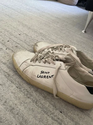 Vita Saint Laurent sneakers - Säljer ett par klassiska vita sneakers från Saint Laurent med handskriven logga på sidan. Skorna har låga skaft, vita snören och beige sula. Tillverkade i läder med stilren och minimalistisk design som passar till det mesta.