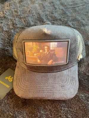 Svart keps från Maggiore med mesh - Säljer en svart keps från Maggiore med mesh på sidorna och bak. Framtill finns en stor patch med konstmotiv och Maggiore-logga. Kepsen har böjd skärm och justerbar snapback. Materialet är en mix av tyg och mesh för en luftig känsla.
