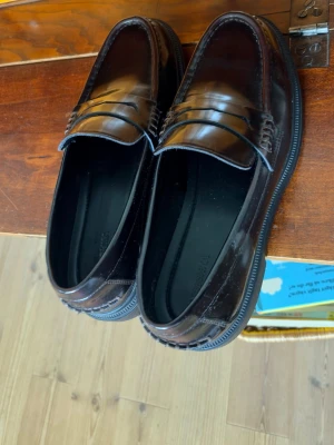 Mörkbruna klassiska loafers i skinn - Säljer ett par mörkbruna loafers i blankt skinn med klassisk pennyloafers-design. Skorna har rundad tå, diskreta sömmar och svart sula. Perfekta för dig som gillar stilrena och tidlösa skor.