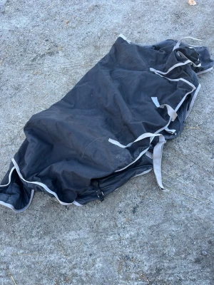 Svart hästtäcke med gråa remmar - Svart hästtäcke med gråa remmar och kantband. Täcket är tillverkat i slitstarkt tyg och har flera spännen och fästen för att sitta bra på hästen. Perfekt för att skydda mot väder och vind storlek 155 och 300 gram.