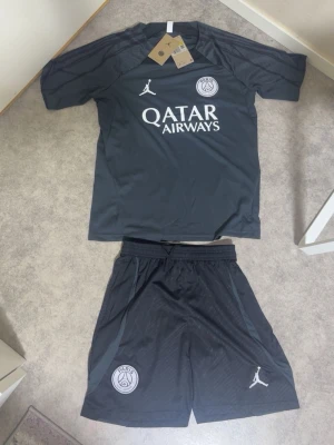 PSG träningsset svart Jordan - Snyggt svart träningsset från PSG x Jordan med t-shirt och shorts. Setet har vita loggor och Qatar Airways-tryck på bröstet. Tillverkat i lätt och ventilerande material, perfekt för fotboll eller träning. Elastisk midja på shortsen och klubbmärke på både tröja och shorts.