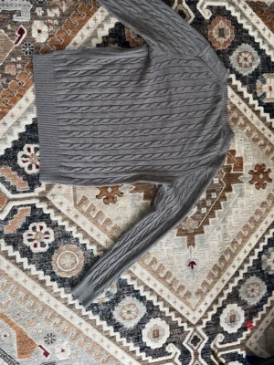 Grå kabelstickad tröja från Gant - Snygg grå kabelstickad tröja från Gant i premium bomull. Tröjan har rund hals, långa ärmar och ribbade muddar vid ärmslut och nederkant. Perfekt för dig som gillar klassisk och stilren look.