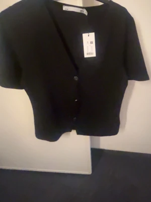 Svart kortärmad kofta med knappar - Stilren svart kofta från Monki med korta ärmar och v-ringad hals. Koftan har en croppad passform och stängs med tre knappar framtill. Perfekt att styla över ett linne eller t-shirt för en enkel men snygg look.