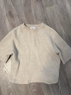 Beige stickad tröja & Other Stories - Kortärmad stickad tröja i mjuk beige ullmix från & Other Stories. Tröjan har rund halsringning med ribbstickad kant men tyvärr ett litet hål, det syns knappt.