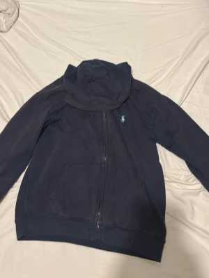 Mörkblå hoodie från Polo Ralph Lauren - Mörkblå hoodie från Polo Ralph Lauren i custom fit med dragkedja framtill och klassisk huva. Har fickor på sidorna och den ikoniska broderade loggan i ljusblått på bröstet. Tillverkad i mjukt bomullsmaterial som känns skönt mot huden tröjan är lite blekt och lite liten i storleken.