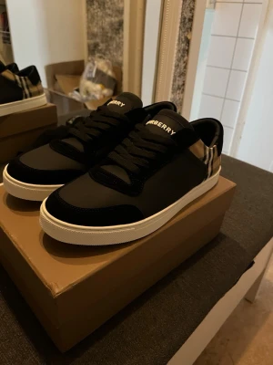 Burberry svart-beige rutiga sneakers - Snygga sneakers från Burberry med klassiskt beige, svart och rött rutmönster på sidan och svart läder samt mockadetaljer. Vita sulor och svarta skosnören. Perfekta för dig som vill ha en stilren och lyxig look med ikoniskt mönster.
