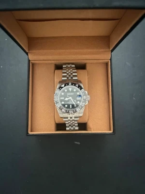 Seiko  - Stilren Seiko gmt herrklocka med svart urtavla, datumvisning och silverfärgad länk i rostfritt stål. Klockan har en svart och silver vridbar bezel med 24-timmarsmarkeringar och tydliga index. Perfekt accessoar för dig som gillar klassisk och sportig stil.