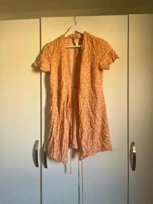 Blommig omlottklänning i orange - Söt kort omlottklänning med småblommigt mönster i orange och vitt. Klänningen har korta ärmar med volangkant och knytband i midjan.  