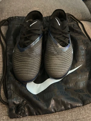 Nike svarta fotbollsskor med blå detaljer - Snygga fotbollsskor från Nike i svart med mörkblå och vita detaljer. Skorna har en stickad ovandel för bra passform och snörning framtill. Swoosh-logga på sidan och tungan. Levereras med svart Nike-påse. Perfekta för dig som vill ha stilrena och funktionella skor på planen.